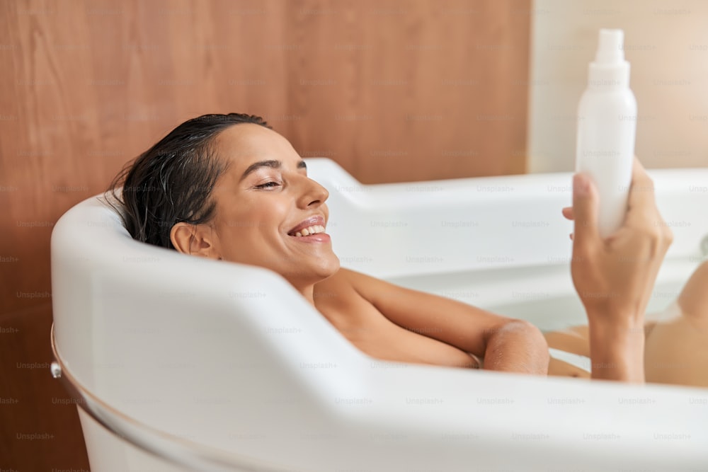 Atractiva dama con el pelo mojado mirando el producto cosmético y sonriendo mientras se relaja en la bañera