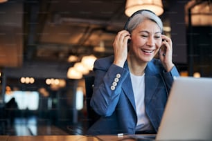 Cheerful businesswoman in headphones with microphone looking at notebook display and smiling while sitting at the table in office