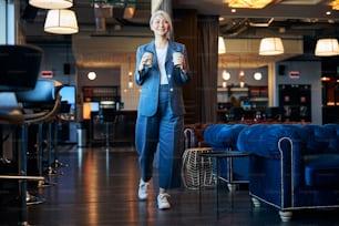 Beautiful lady holding two takeout coffee cups and smiling while strolling down the cafeteria