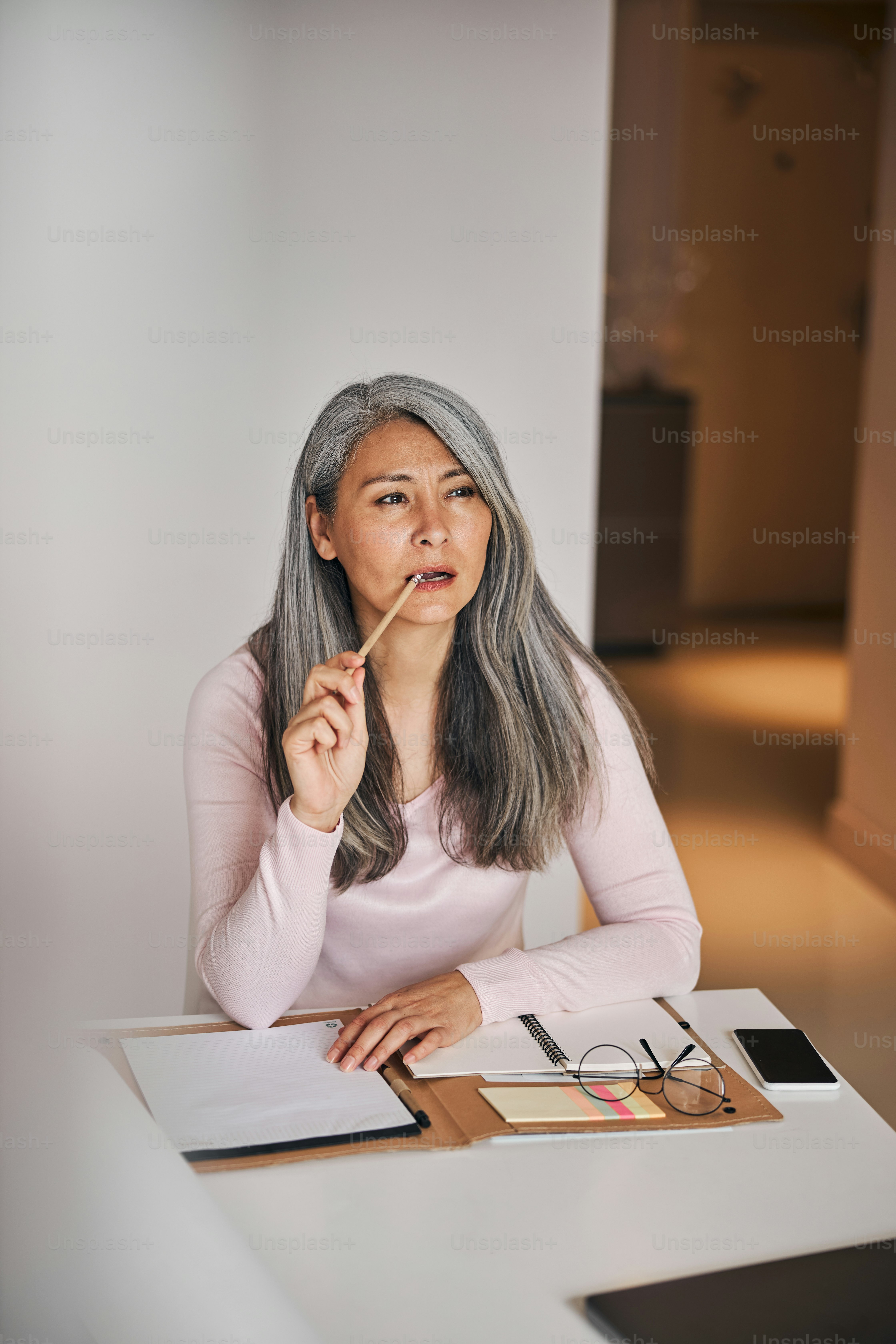 매력적인 아가씨는 종이와 연필을 물고 테이블에 앉아 프로젝트에 대한 최고의 아이디어에 대해 숙고하고 있습니다.