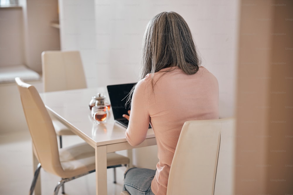 Back view of woman sitting at the table with cup of tea and using modern laptop