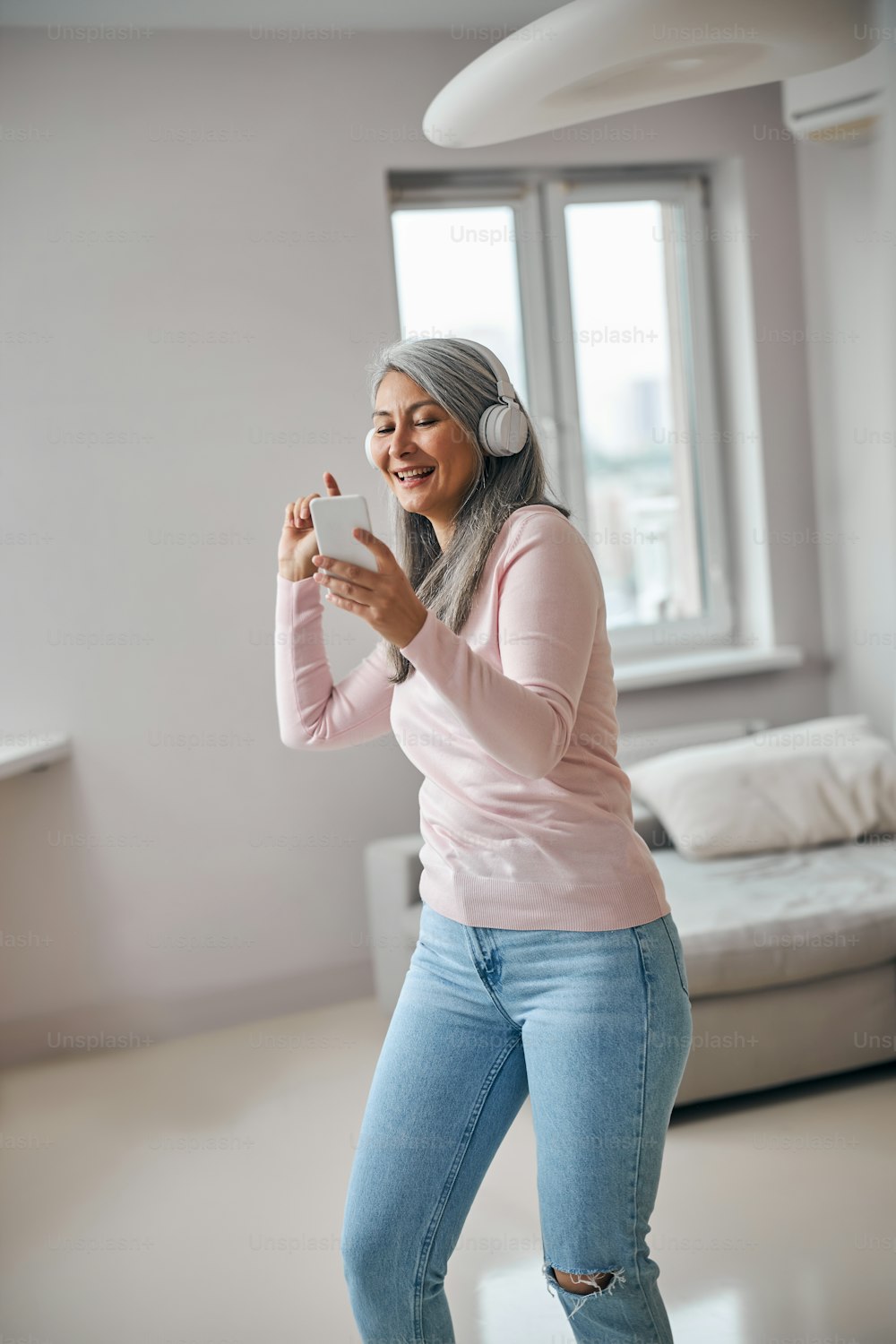 Cheerful lady in wireless headphones holding cellphone and smiling while moving to the rhythm of music in apartment