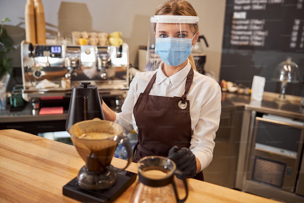 Retrato de primer plano de una mujer camarero con máscara protectora y guantes durante la epidemia de coronavirus que prepara café mientras usa una tetera de goteo