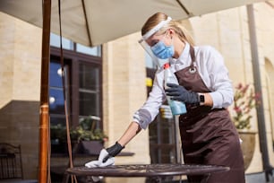 Side view portrait of young beautiful woman waiter cleaning table with disinfectant in cafe during coronavirus outbreak