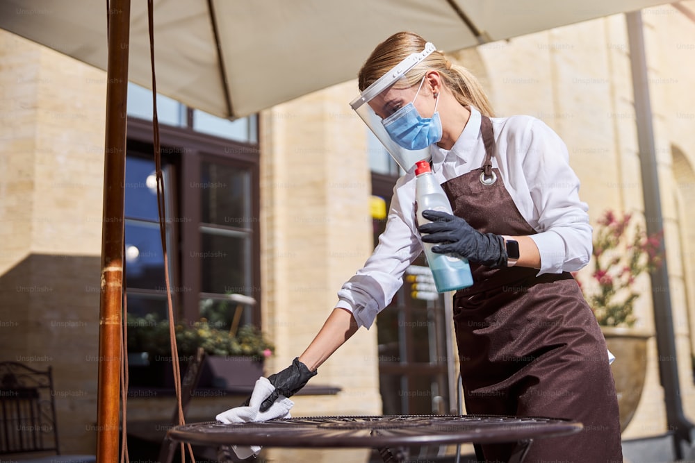 Side view portrait of young beautiful woman waiter cleaning table with disinfectant in cafe during coronavirus outbreak