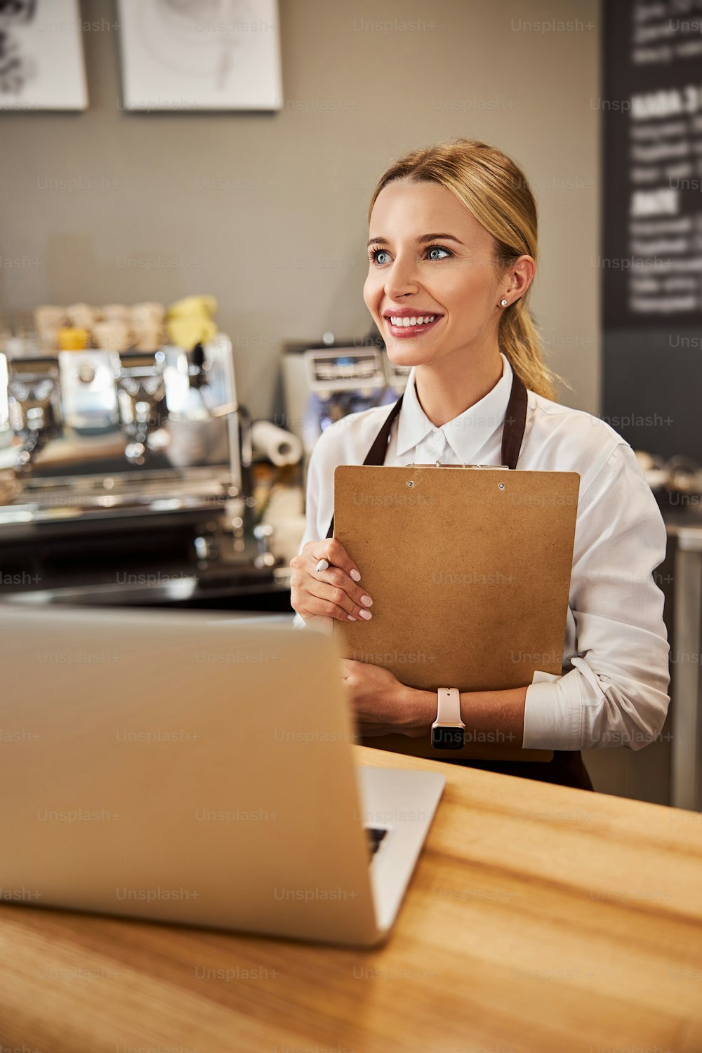 Close up portrait of happy cheerful female with clipboard in hands looking away while standing near the wooden table