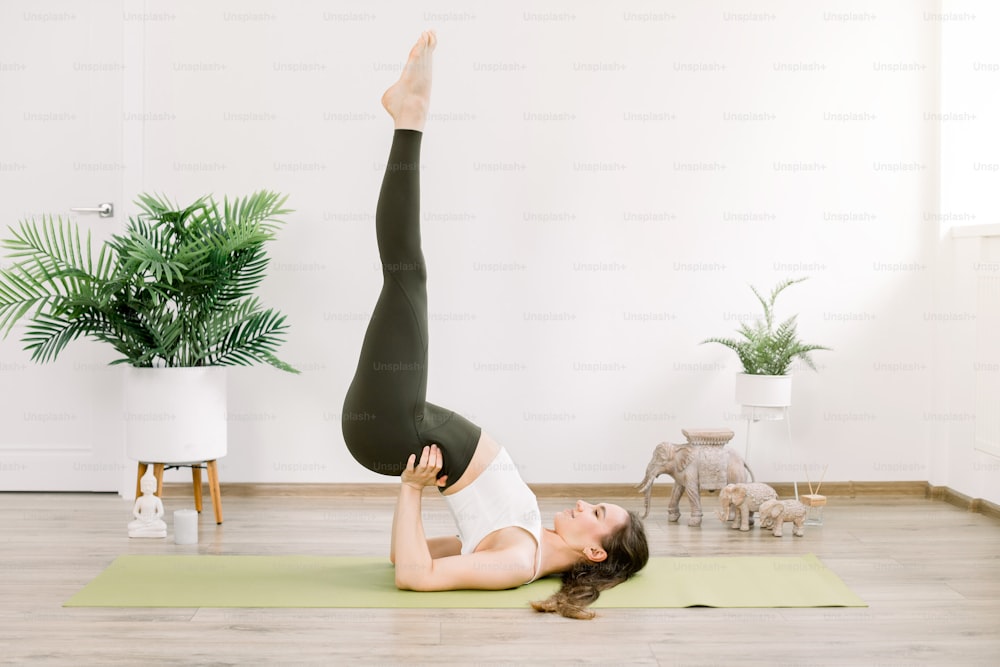 Side full length view of young european woman practicing yoga, standing in Viparita Karani exercise, shoulderstand pose, working out on green mat at cozy fit studio.