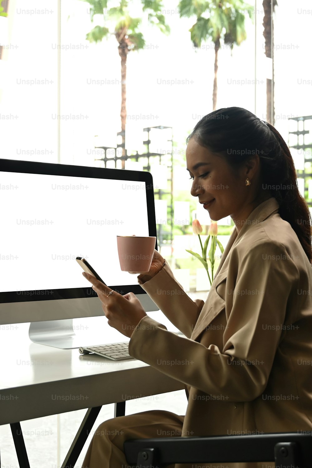 A young Asian businesswoman working on computer and reading incoming notification on smartphone at office.