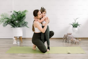 Mother and daughter spending time together doing yoga exercise home. Pretty woman and little child girl, hugging each other after joint fitness training. Family sport concept.