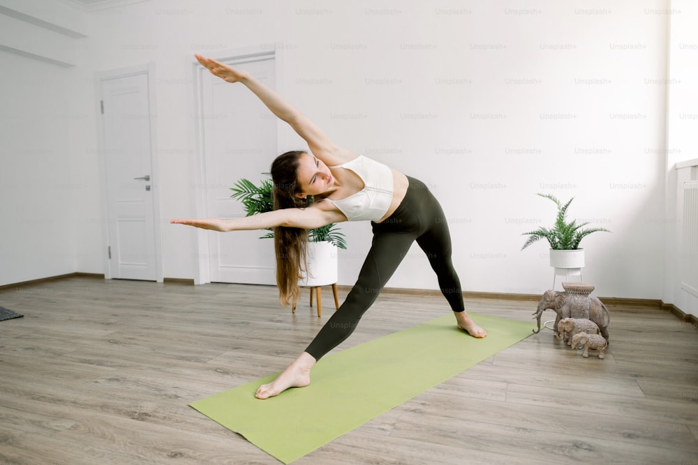 Prise de vue horizontale en intérieur d’un joli jeune canapé de yoga de fitness, debout dans une pose de yoga en triangle flottant sur un tapis vert, profitant de son temps sportif le matin.