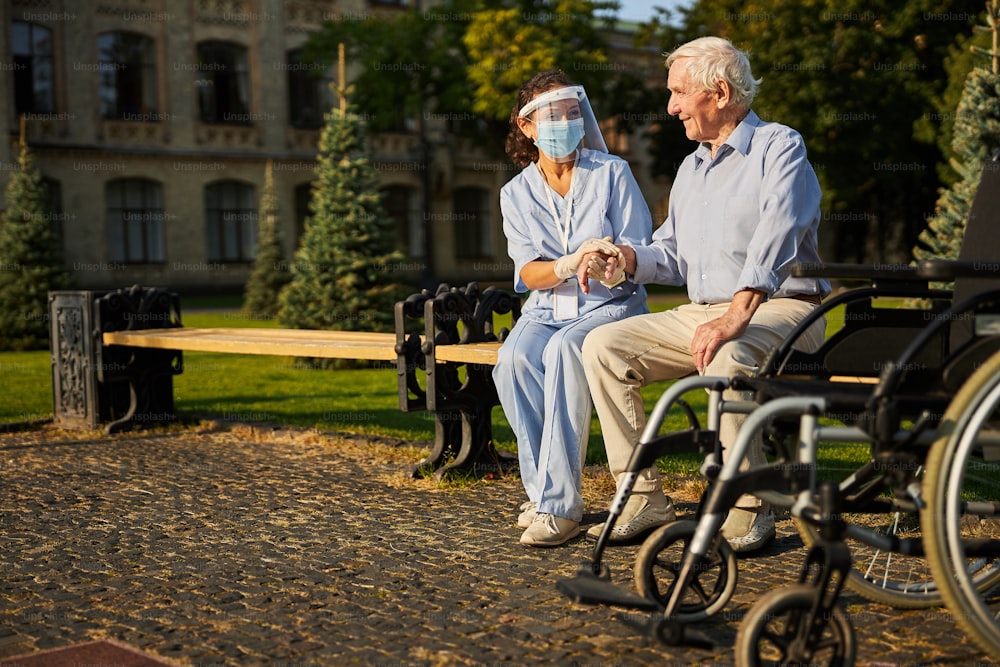 Happy patient sitting on the bench and holding caregiver for a hand while spending time together outside