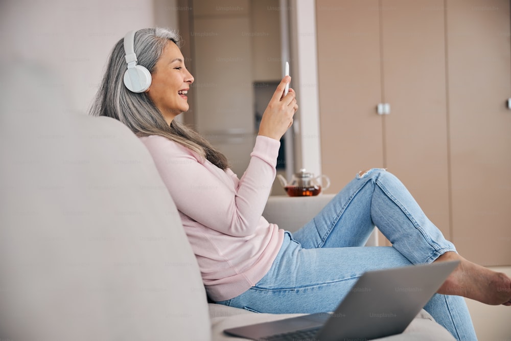 Ritratto di vista laterale di donna allegra felice in abiti casual che guarda lo schermo il suo telefono cellulare e ride nel soggiorno