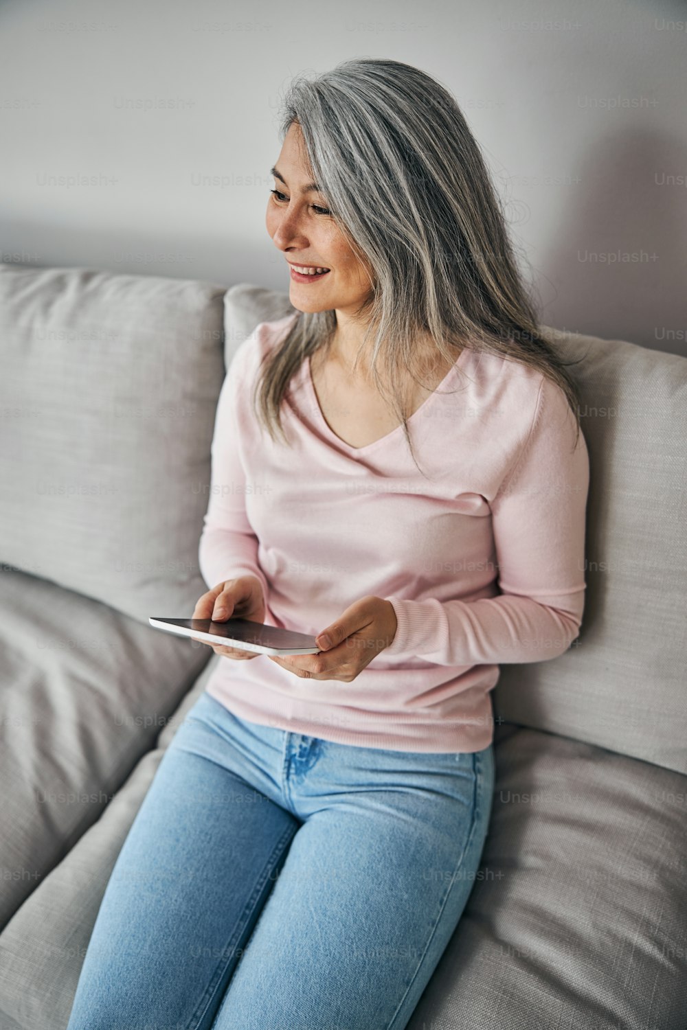 Top view portrait of happy smiling pretty lady in casual clothes holding tablet in hands while sitting on the grey sofa in room indoors