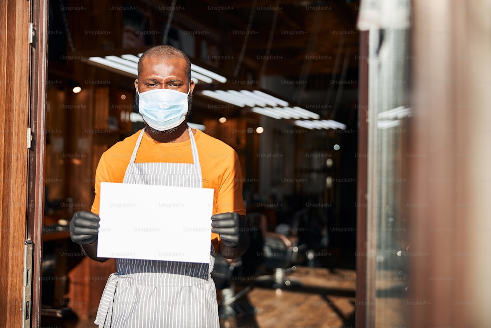 Handsome Afro American man with blank mockup placard in his hands wearing protective face mask, sterile gloves and apron