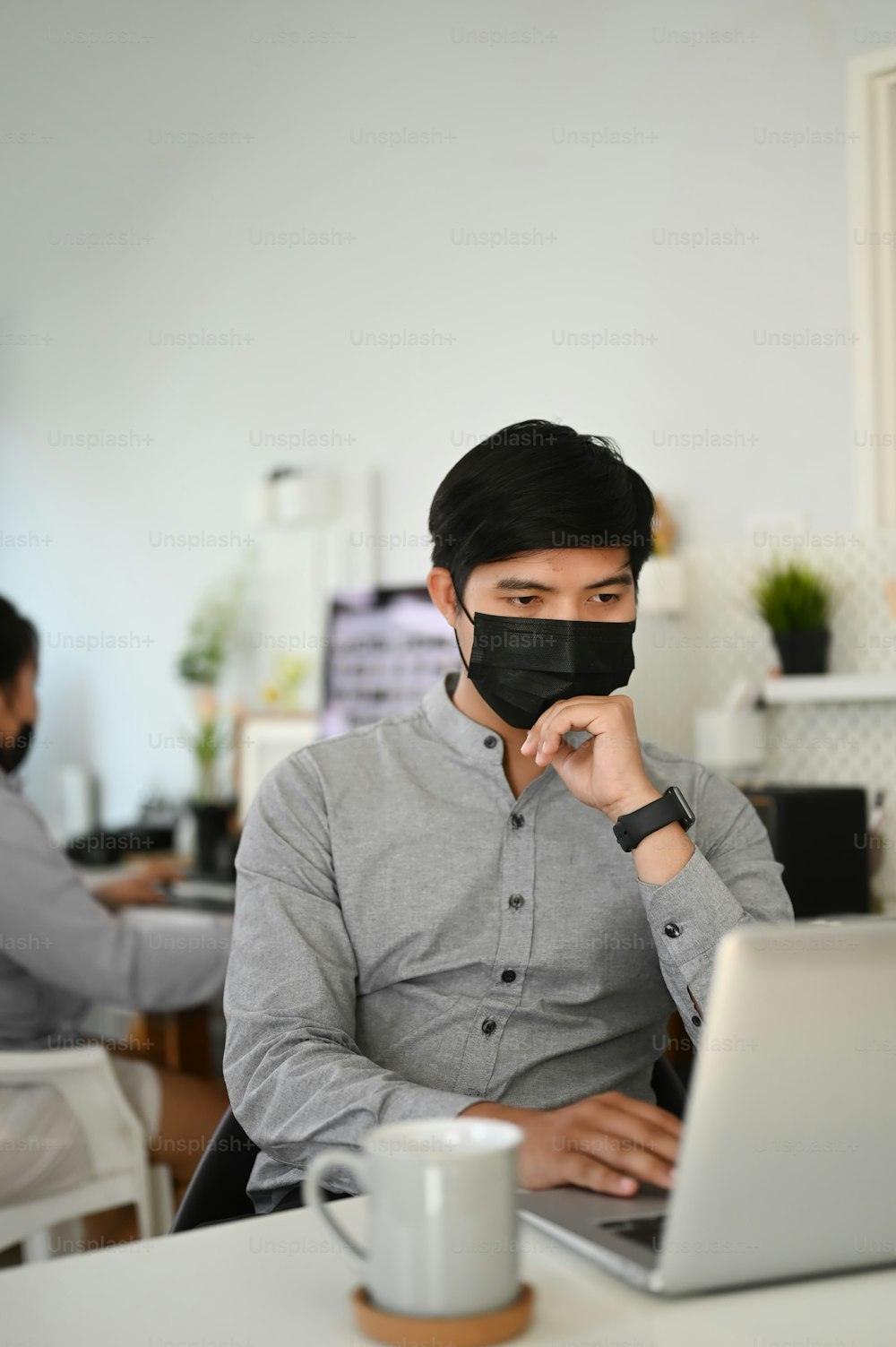 Porträt eines männlichen Freelancer-Fotografen mit medizinischer Gesichtsmaske, der in seinem Büro am Laptop arbeitet.