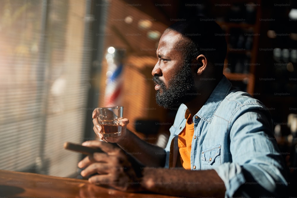 Handsome serious gentleman holding cigar and glass of alcoholic drink while sitting at the bar counter