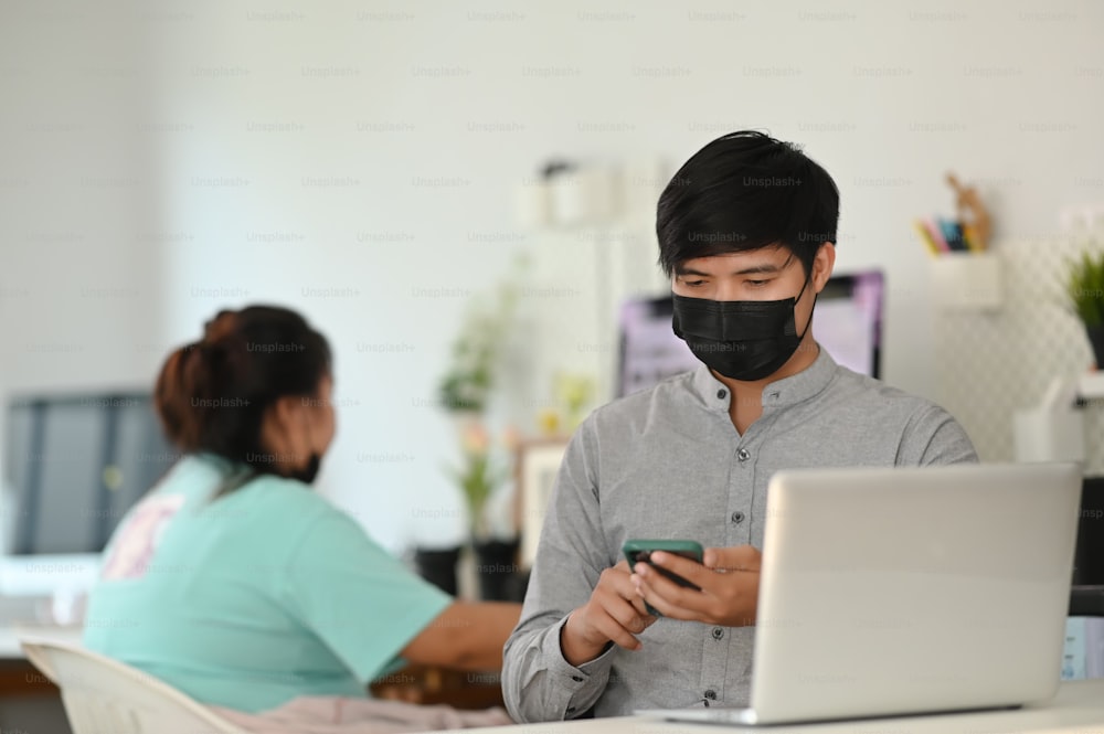 Ein junger Mann mit Gesichtsmaske mit Mock-up-Smartphone, während er vor einem Laptop in einem komfortablen Arbeitsbereich sitzt.