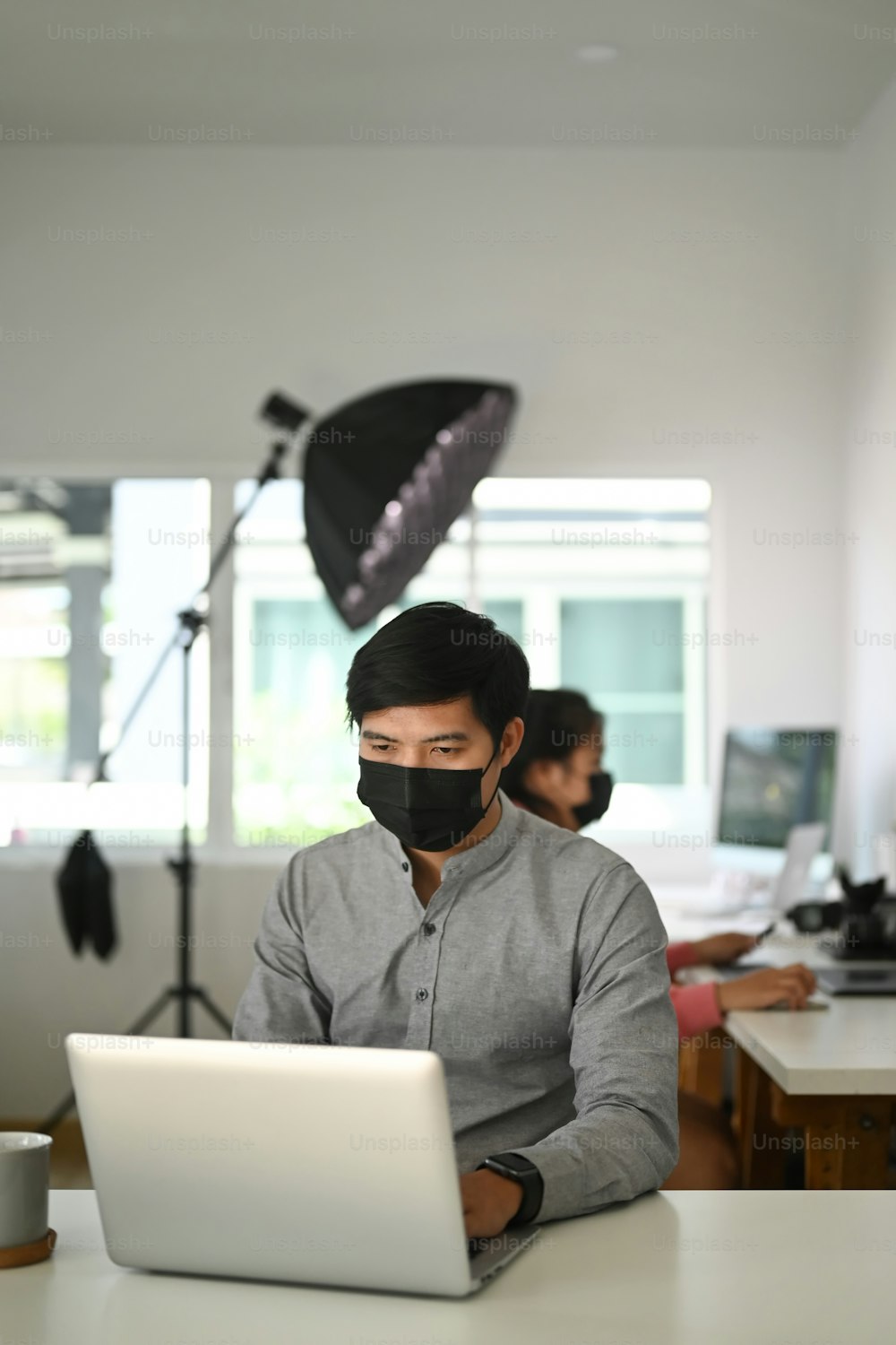 Porträt eines männlichen Freiberuflerfotografen mit Gesichtsmaske überprüft Fotos auf einem Laptop, während er am Tisch am Arbeitsplatz sitzt.