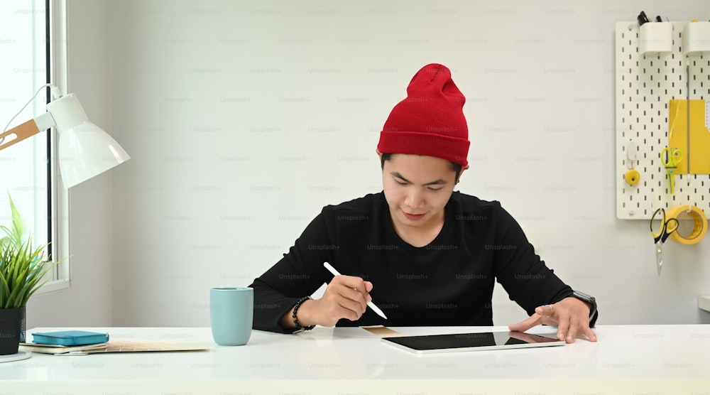 A Graphic designer man in red wool hat retoucher working with tablet and stylus pen in studio.