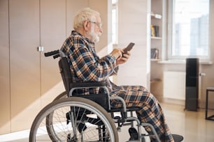 Vue latérale d’une personne âgée assise dans un fauteuil roulant avec un téléphone portable dans les mains
