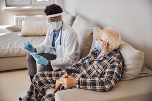 Médico de pelo oscuro con un protector facial sentado en la cama junto a un anciano