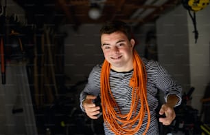 Portrait of down syndrome adult man with rolled up cable around neck in shed looking at camera.