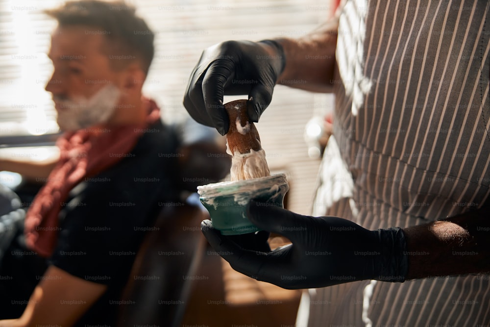 Close up of man hairdresser hands in sterile gloves dipping shave brush into bowl with cream while gentleman sitting in barber chair