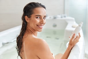 Beautiful lady with cosmetic product in her hand looking at camera and smiling while relaxing in bathtub