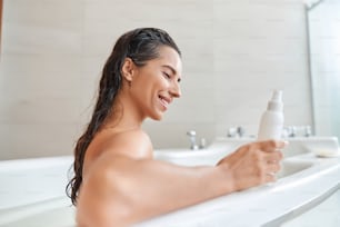 Beautiful lady with wet hair looking at cosmetic product and smiling while taking bath