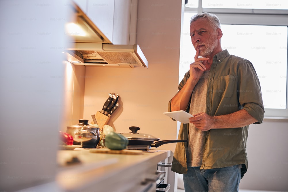 Grey-haired gentleman touching chin and looking thoughtful whille standing in kitchen with a tablet