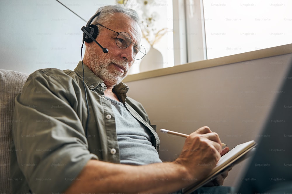Anciano experimentado que usa anteojos y auriculares mientras toma notas y mira la computadora