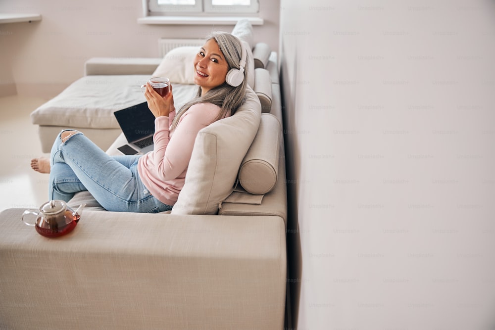 Back view portrait of charming adult female in pink sweater looking at the photo camera while drinking tea while spending time on sofa in flat