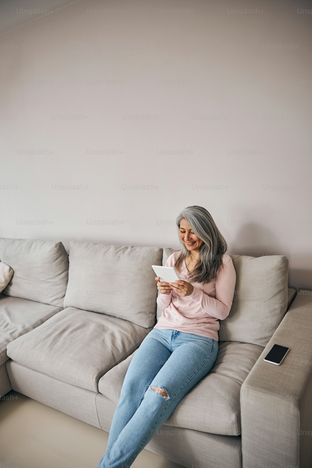 Porträt einer glücklich lächelnden Frau, die auf dem grauen Sofa sitzt, während sie ein modernes Gerät im Wohnzimmer verwendet