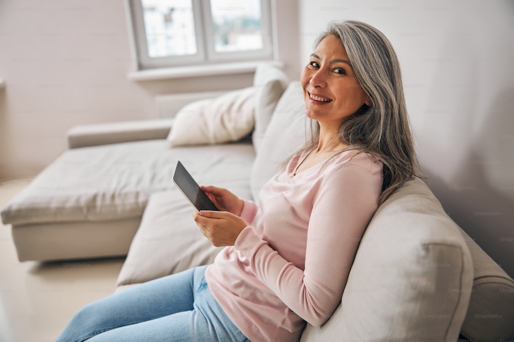 Side view portrait of beautiful Asian adult woman in Casula clothes holding modern tablet in hands while sitting on the living room
