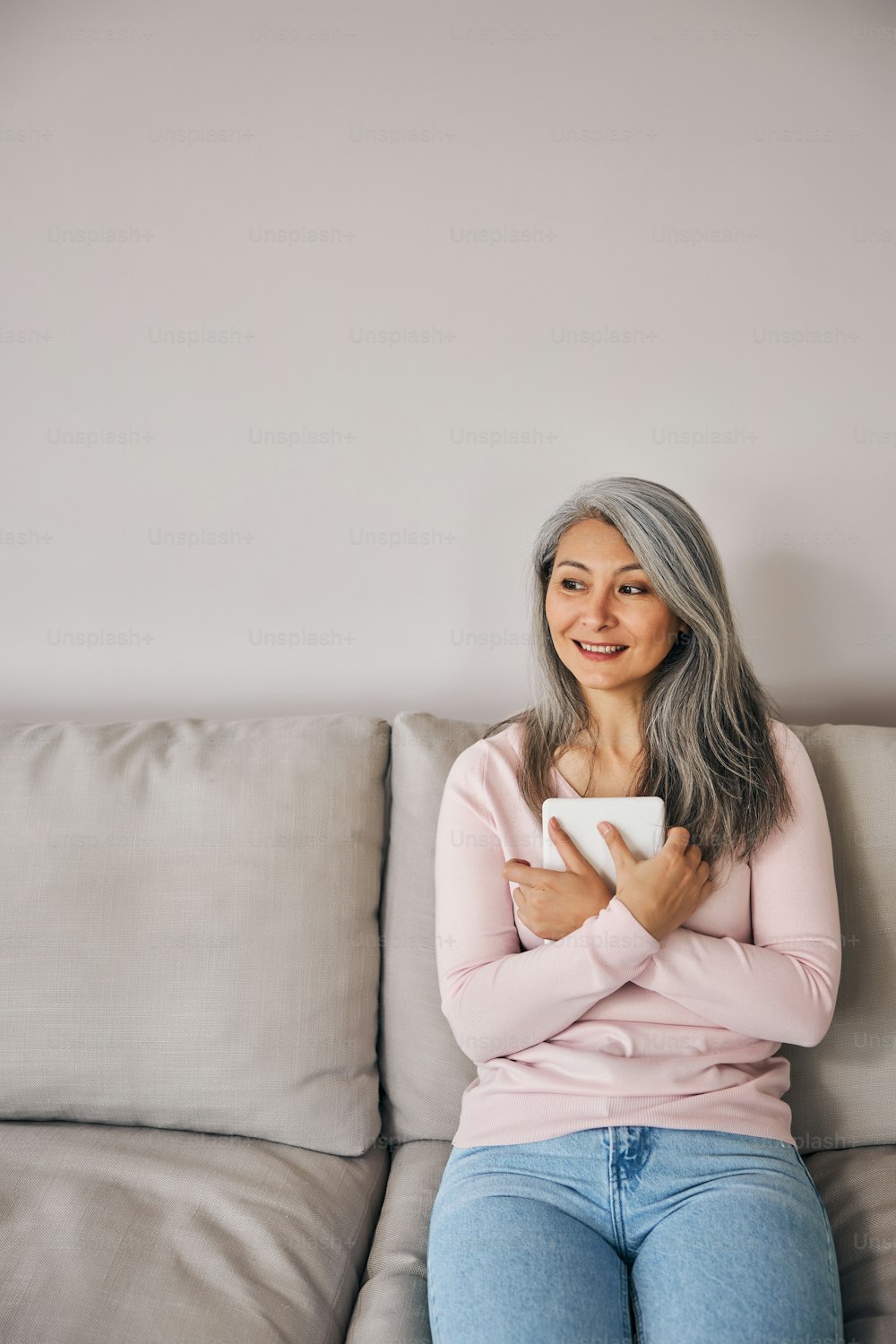 Close up portrait of charming elegant woman in pink sweater and jeans sitting on gray sofa while crossing hands with tablet in front of herself