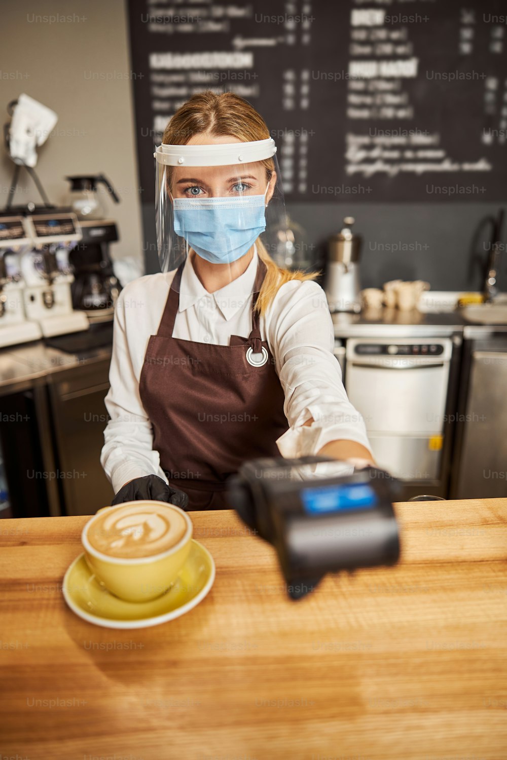 Close up portrait of saleswoman in medical mask standing near the wooden table in cafe with payment machine in hand