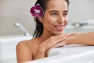 Primo piano di bella signora con orchidea viola nei suoi capelli bagnati che distoglie lo sguardo e sorride mentre si rilassa nella vasca da bagno