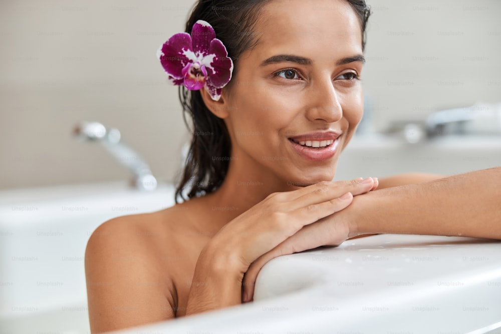 Primer plano de una dama bonita con orquídea violeta en su cabello mojado mirando hacia otro lado y sonriendo mientras se relaja en la bañera