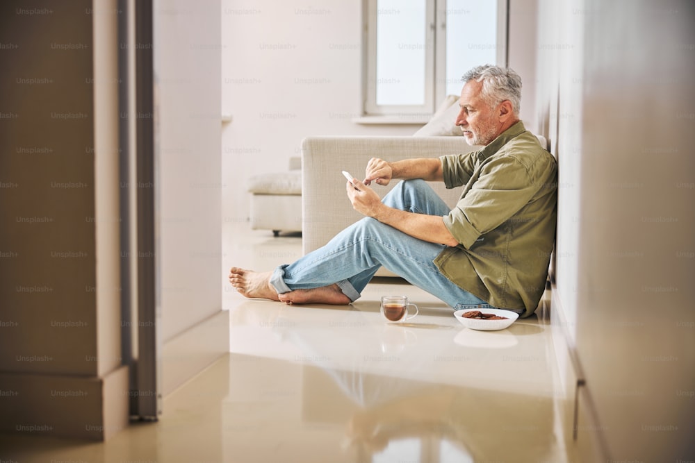 Anziano signore nei suoi abiti casual che fa clic sul suo smartphone mentre è seduto sul pavimento con una tazza di tè