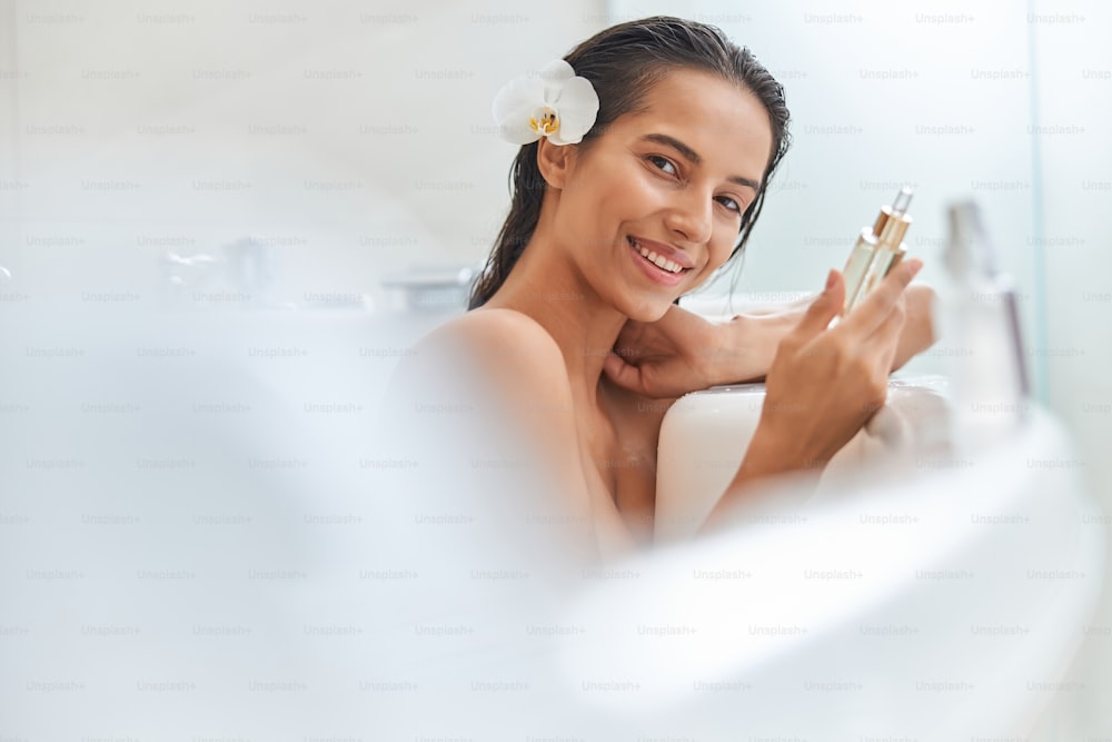 Bella signora con orchidea bianca tra i capelli che tiene una bottiglia di olio essenziale e sorride mentre si rilassa nella vasca da bagno