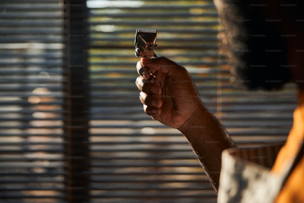 Close up of Afro American man with metal hair clipper in his hand standing by the window with blinds