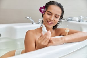 Bella signora con fiore viola nei capelli bagnati che guarda il prodotto per la cura della pelle e sorride mentre si rilassa nella vasca da bagno