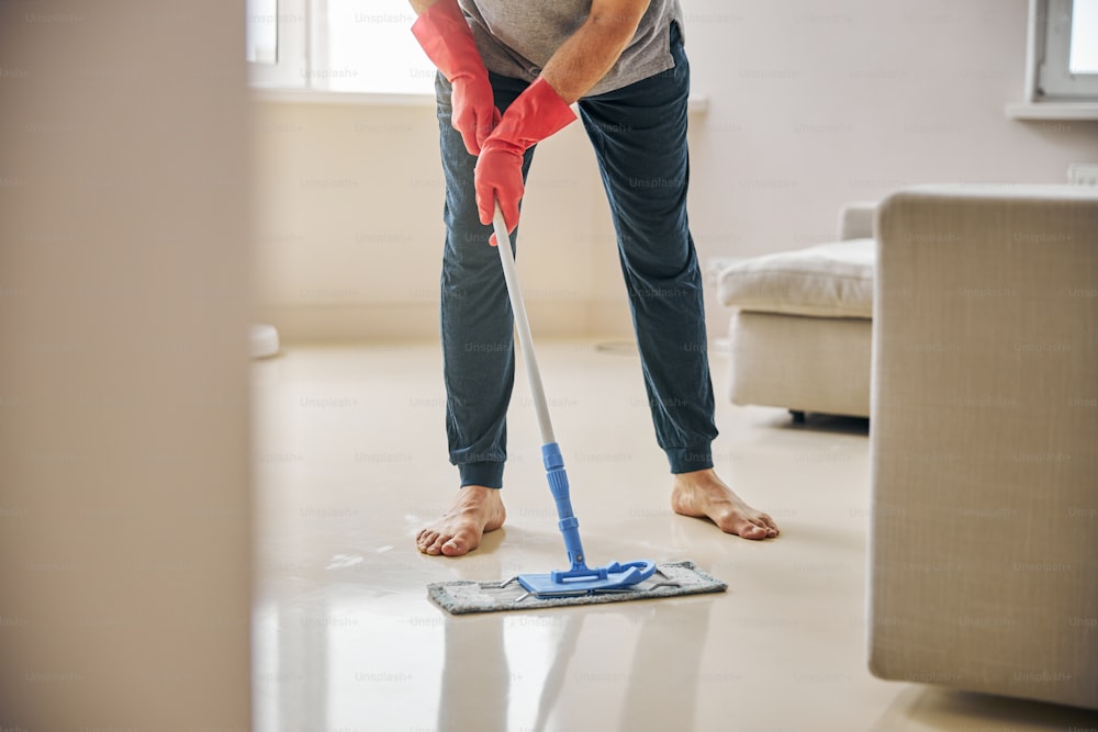 Neat man in rubber gloves standing barefoot while wiping the floor with a wet mop