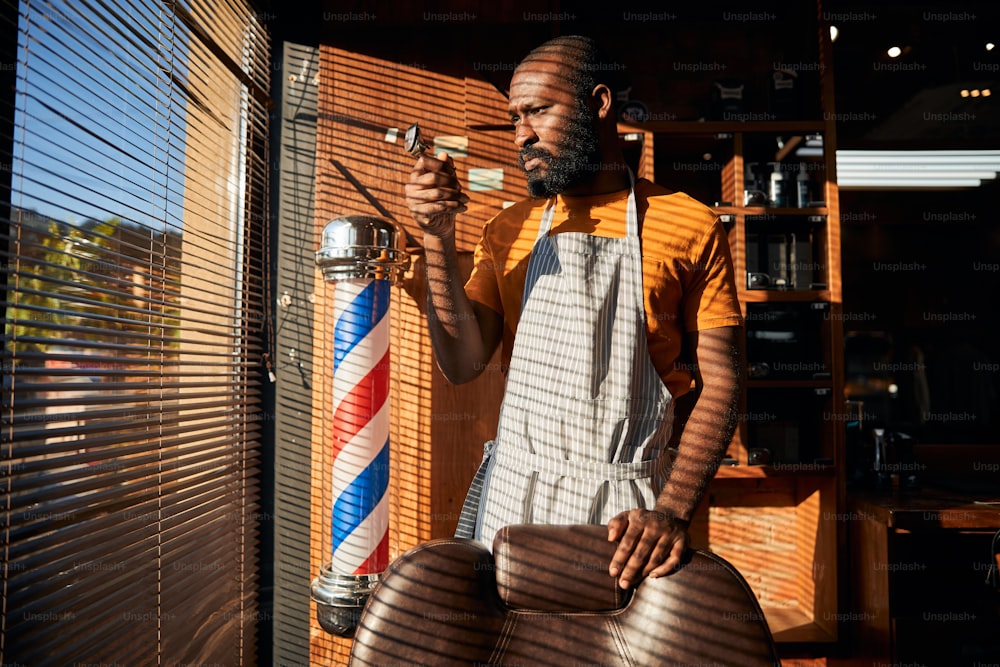 Bearded Afro American man looking at hair clipper in his hand with serious expression while standing by the window in barbershop
