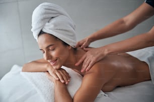 Beautiful lady with towel on her head lying on massage table and smiling while receiving therapeutic back massage