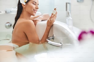 Beautiful lady with flower in her hair looking at skincare product and smiling while relaxing in bathtub