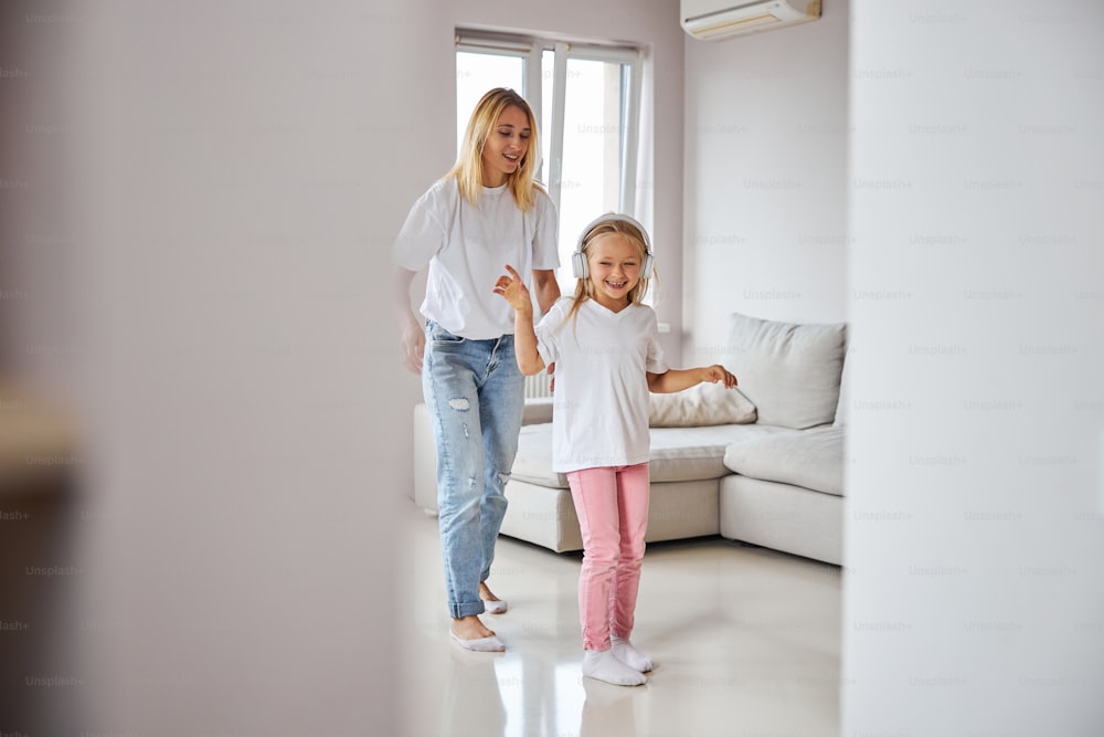 Full length portrait of little girl standing in front of her mother while listening music in headphones and dancing together in their apartment