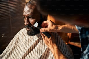 Jovem afro-americano bonito olhando para o lado com expressão séria enquanto barbeiro retirava restolho de sua bochecha com máquina de barbear elétrica