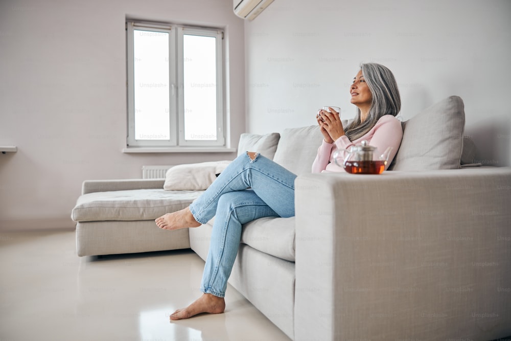 Charming lady holding cup of hot drink and smiling while resting on comfortable sofa in living room