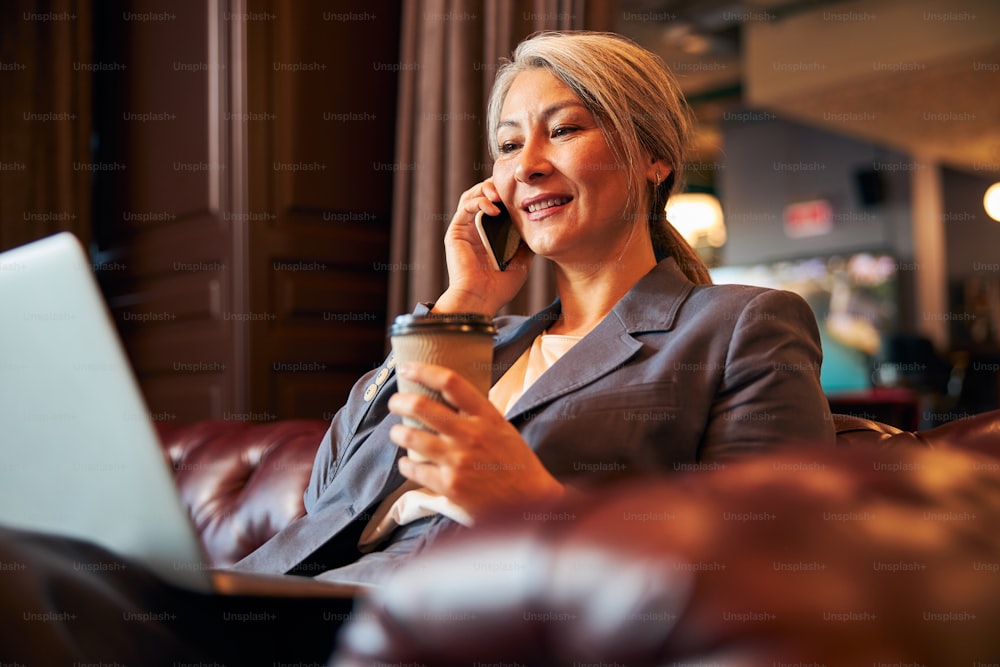 Beautiful woman with cup of coffee in her hand having phone conversation and smiling while working on notebook at business center